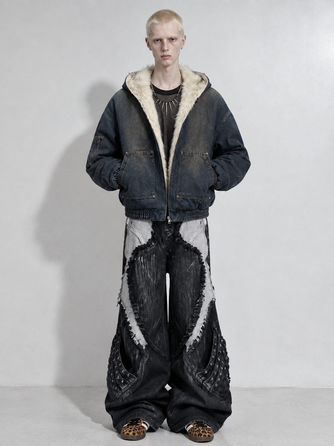 A model stands against a light gray studio background wearing a dark washed denim jacket with a cream sherpa-lined hood, matching wide-leg distressed denim pants, and leopard-print shoes.